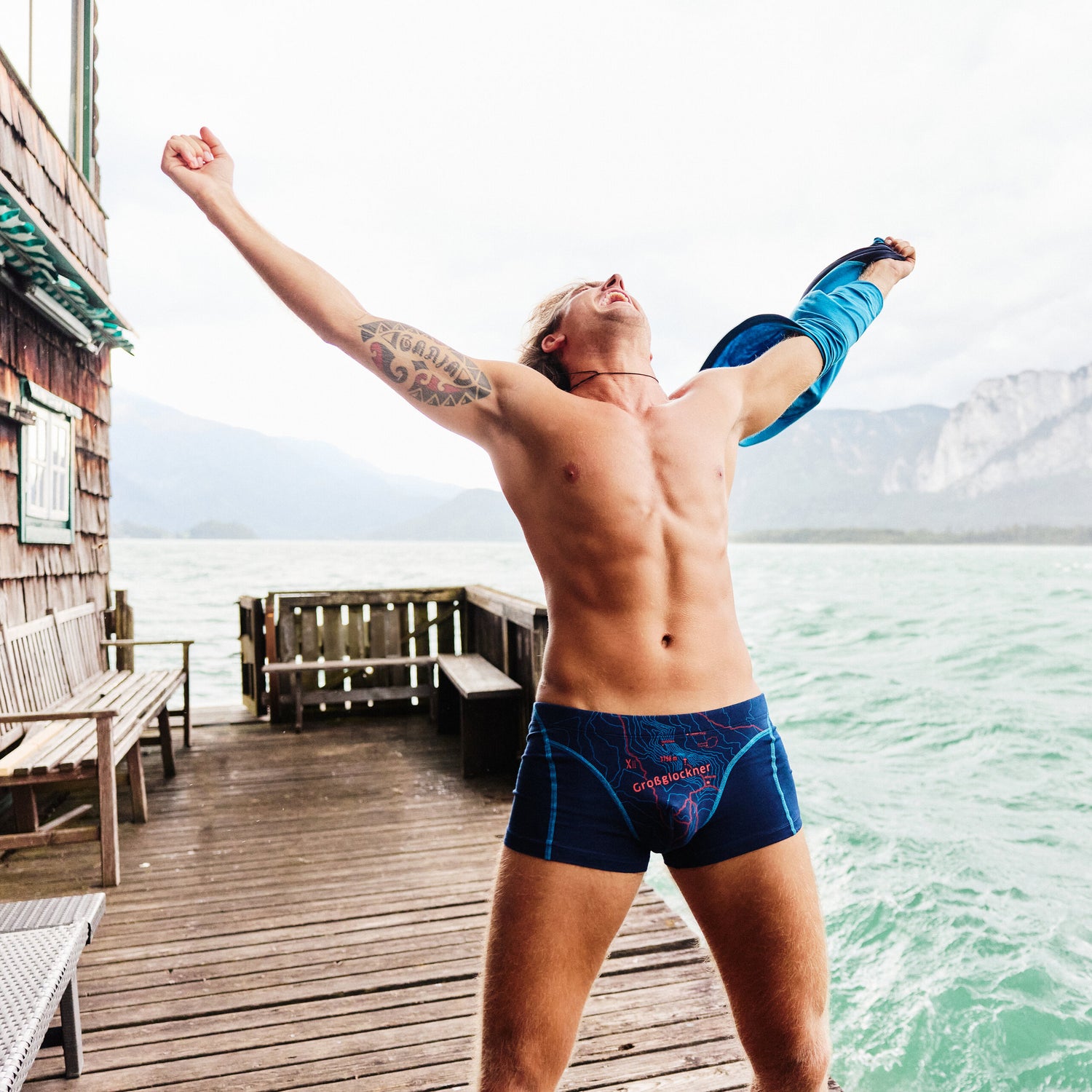 Mann in blauer Boxershorts mit Großglockner-Design, hebt jubelnd die Arme auf einem Steg am See