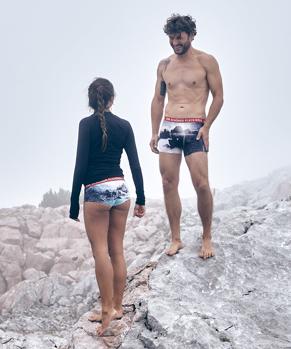 Frau und Mann stehen barfuß auf Felsen in nebeliger Berglandschaft, tragen Unterwäsche mit Landschaftsmotiven und rotem Bund der Marke „Ein schöner Fleck Erde“.