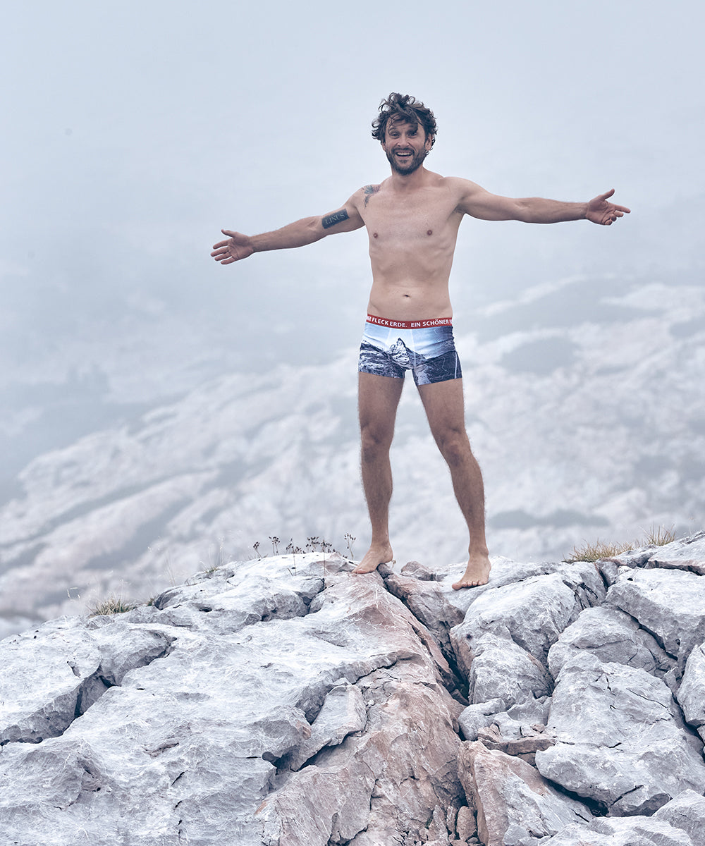 Mann steht barfuß auf Felsen in nebeliger Berglandschaft, breitet die Arme aus und trägt Unterwäsche mit Bergmotiv und rotem Bund der Marke „Ein schöner Fleck Erde“.