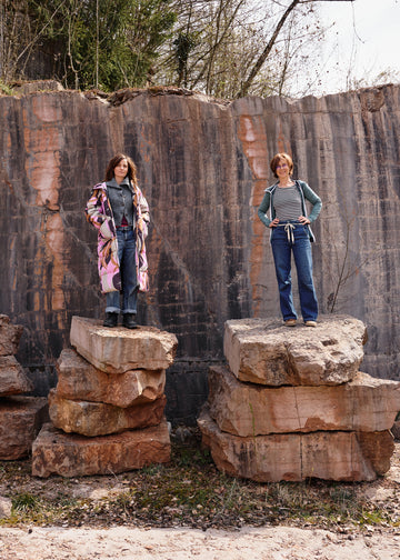 Zwei Personen stehen auf gestapelten Steinblöcken vor einer Felswand; links ein langer, bunt gemusterter Mantel über grauem Strick und Jeans, rechts ein gestreiftes Shirt mit grünem Kapuzenpullover und Jeans.