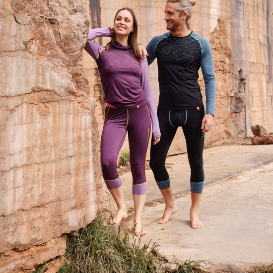 Frau und Mann tragen Funktionswäsche von Ein schöner Fleck Erde – sie in Violett, er in Dunkelblau – und stehen gemeinsam lächelnd vor einer Felswand in der Natur