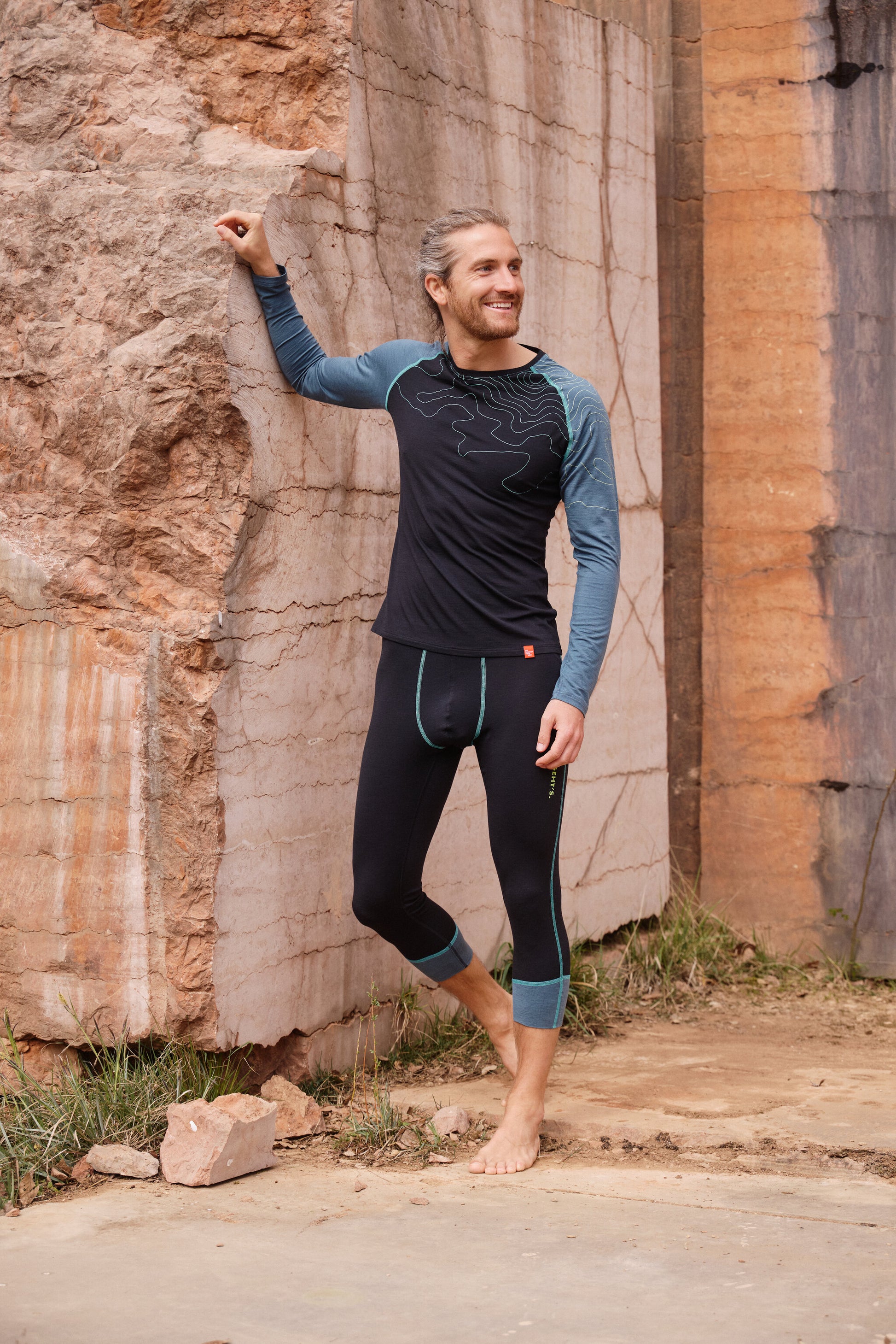 Mann lehnt entspannt an Felswand und trägt dunkelblaue Funktionswäsche mit hellblauen Details von Ein schöner Fleck Erde mit Türkisem Nahtdesign