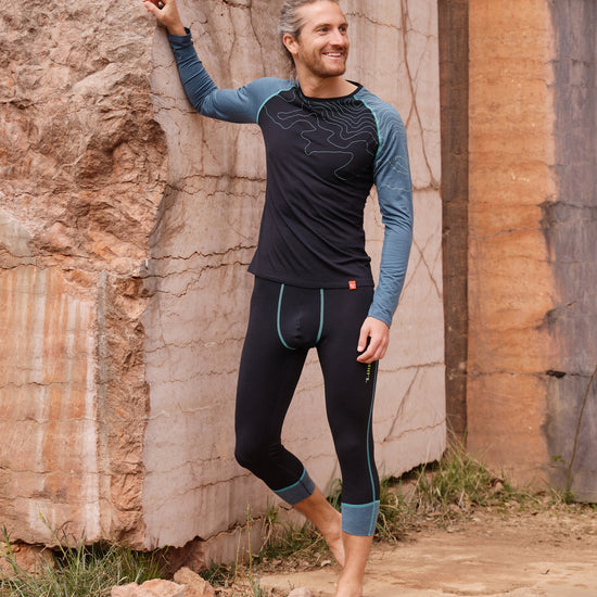 Mann trägt dunkelblaue Funktionswäsche mit hellblauen Ärmeln und Linienmuster von Ein schöner Fleck Erde, steht lächelnd an einer Felswand im Freien