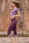 Frau trägt lilafarbenes T-Shirt und dreiviertellange violette Funktionsleggings mit gelben Nähten von Ein schöner Fleck Erde, steht lächelnd vor Felswand im Freien