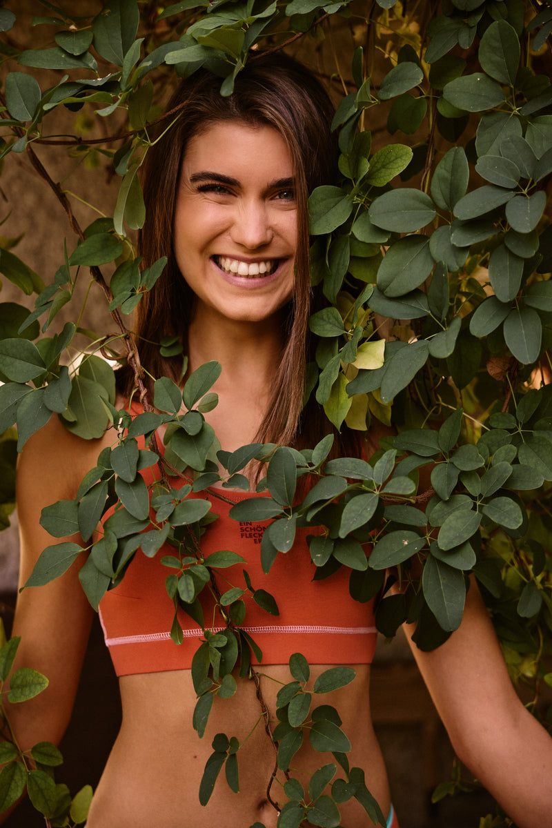 Damen Bustier von Ein schöner Fleck Erde in Orange mit Markenlogo, Nahaufnahme Oberkörper, Model steht hinter grünen Blättern, lächelnde Pose, Outdoor-Fotoshooting bei natürlichem Licht