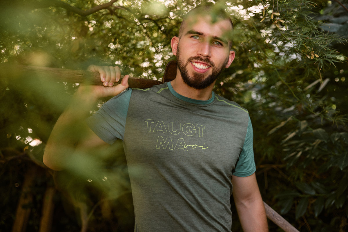 Herren T-Shirt von Ein schöner Fleck Erde in Grün mit gelbem Aufdruck ‚TAUGT MAHOI‘, kurze Ärmel, Nahaufnahme am Oberkörper, Outdoor-Fotoshooting im Garten, Model hält Holzstab über der Schulter, natürliche Pose, Pflanzen im Hintergrund