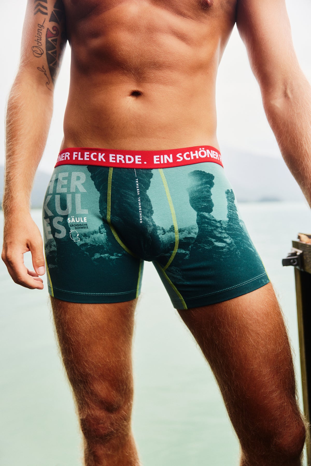 Nahaufnahme Herren Boxershorts von Ein schöner Fleck Erde in Grün mit Felsen-Motiv und Aufdruck ‚HERKULES SÄULE‘, roter elastischer Bund mit Markenlogo, bequemer Schnitt, Outdoor-Fotoshooting am See, Holzsteg im Hintergrund