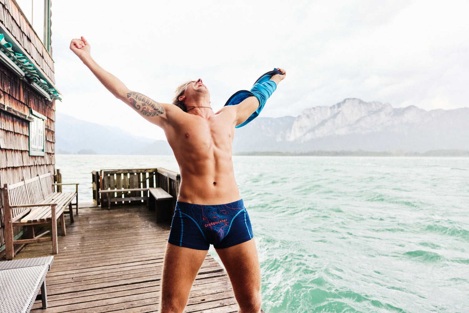 Mann in blauer Unterwäsche mit „Großglockner“-Print steht barfuß auf einem Steg am See und reißt die Arme siegreich in die Luft, mit Bergen im Hintergrund.
