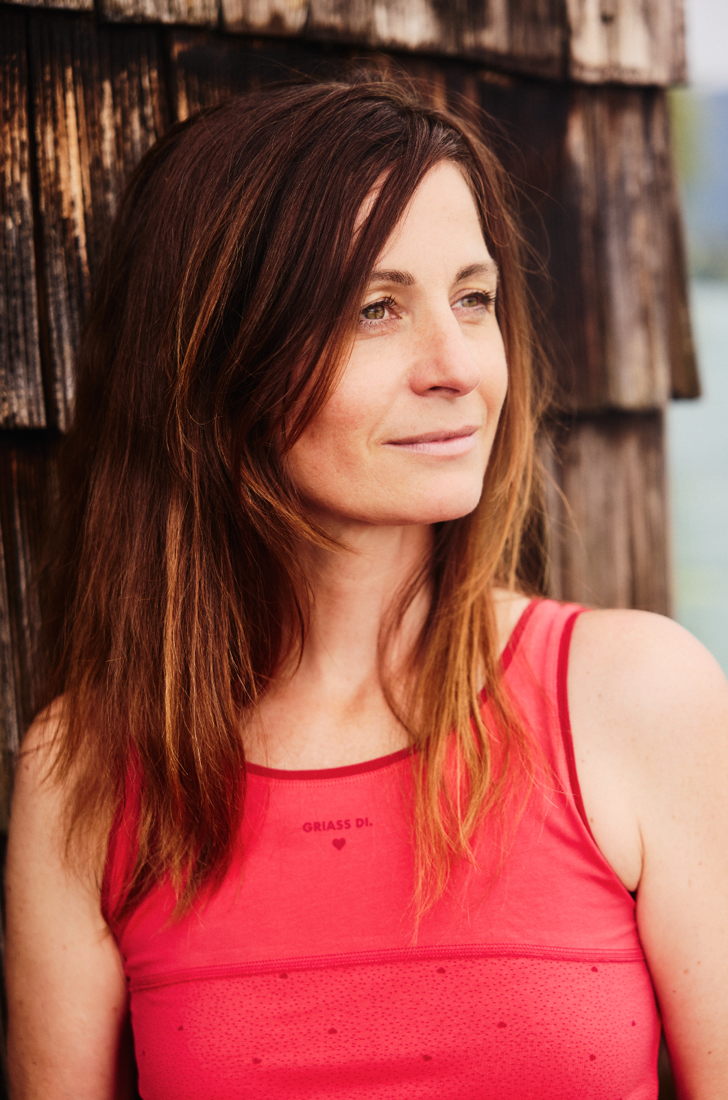 Frau trägt rotes Top mit Aufdruck ‚Griass Di‘ von Ein schöner Fleck Erde vor rustikaler Holzwand