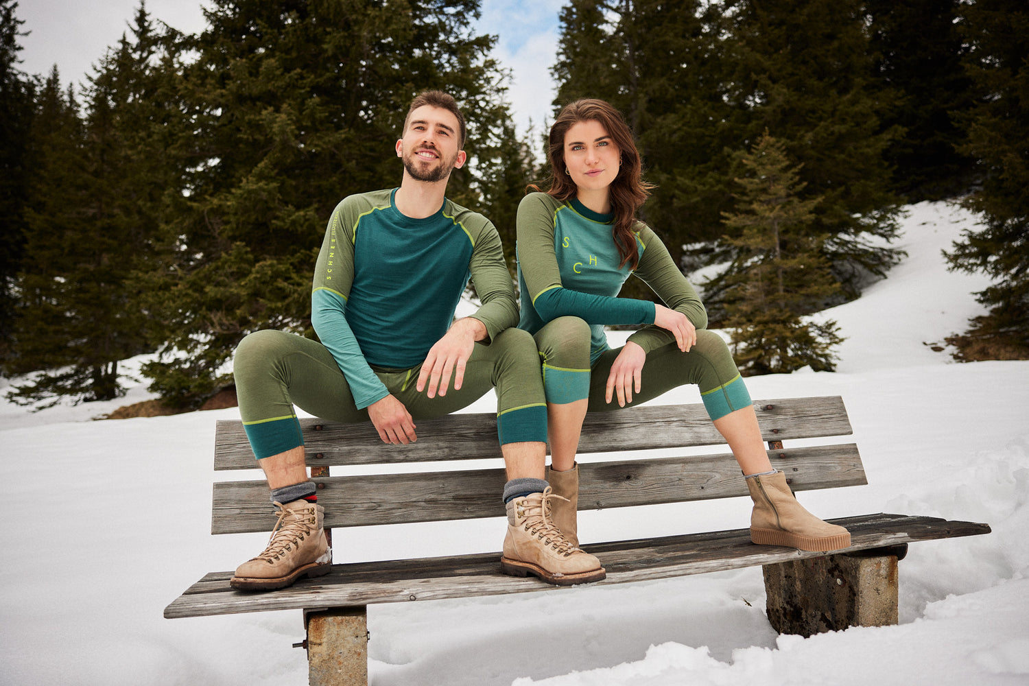 Mann und Frau in grüner Funktionsunterwäsche von Ein schöner Fleck Erde, sitzend auf einer Holzbank im Schnee, Langarmshirt und Leggings-Set, Outdooraufnahme in verschneiter Berglandschaft mit Tannen im Hintergrund