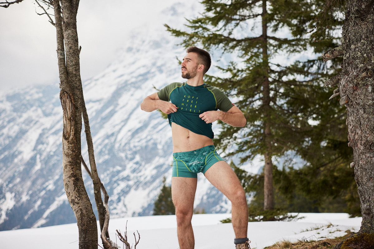 Mann in grüner Unterwäsche mit Bergmotiv von Ein schöner Fleck Erde posiert im Schnee vor alpiner Bergkulisse