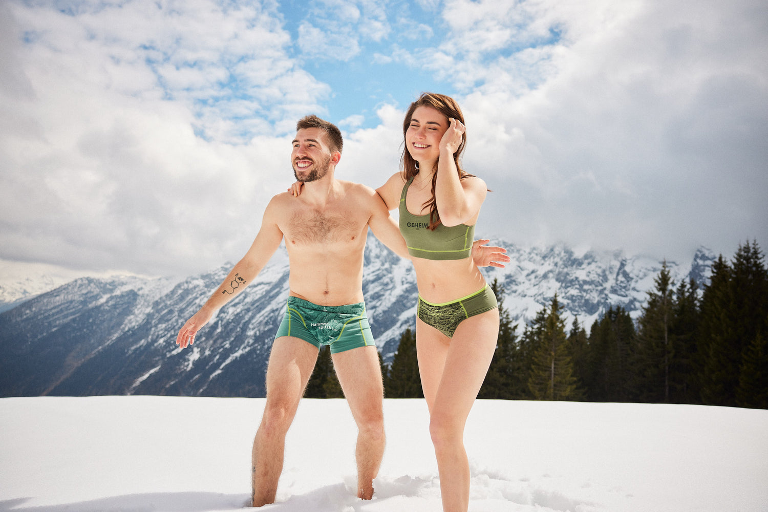 Paar in grüner Unterwäsche mit Bergmotiven von Ein schöner Fleck Erde steht lachend im Schnee vor alpiner Berglandschaft