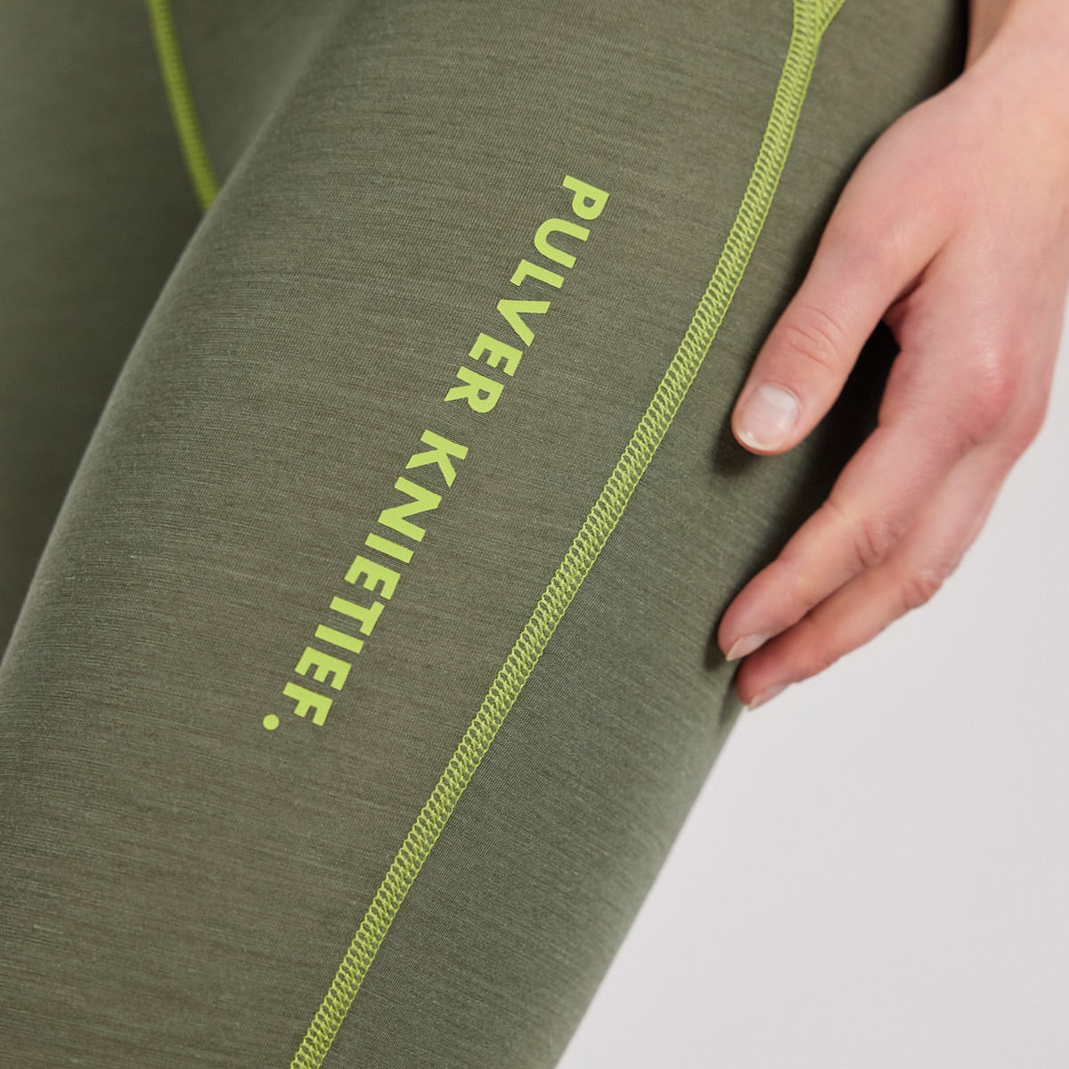 Damen Funktionshose von Ein schöner Fleck Erde in Olivgrün mit gelber Naht und Aufdruck ‚PULVER KNIETIEF‘, Nahaufnahme am Bein, Teil des Funktionswäsche-Sets, Indoor-Fotoshooting, natürliche Pose