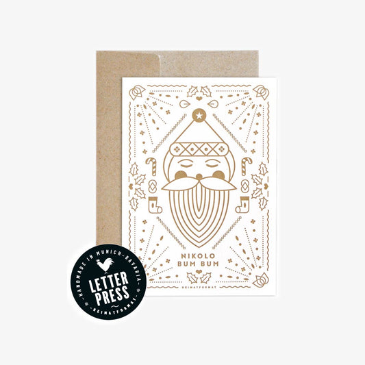 Weihnachtliche Grußkarte mit geprägter Illustration eines stilisierten Nikolausgesichts und der Aufschrift „Nikolo Bum Bum“, beigelegt in einem braunen Umschlag. Links unten ein rundes Siegel mit der Aufschrift „Handmade in Munich – Bavaria | Letterpress | Heimatformat“.